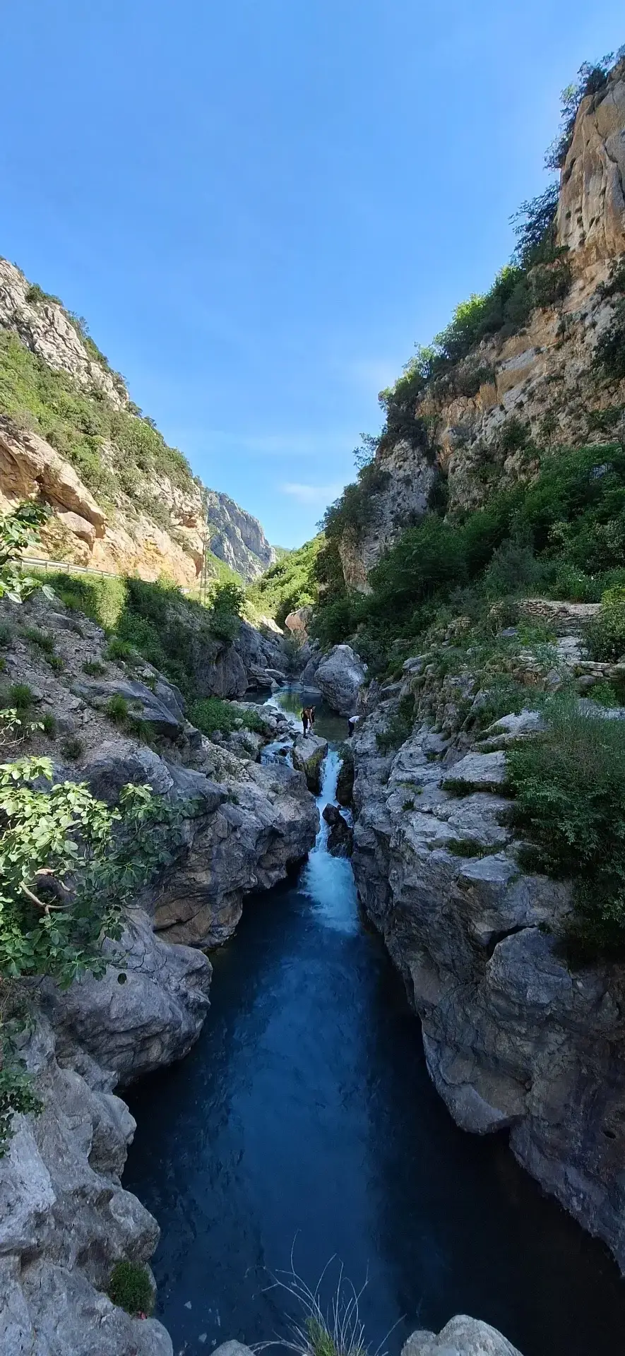 A small waterfall drops into a deep blue pool inside Bovilla Canyon, a narrow limestone gorge with two people standing at the water's edge.