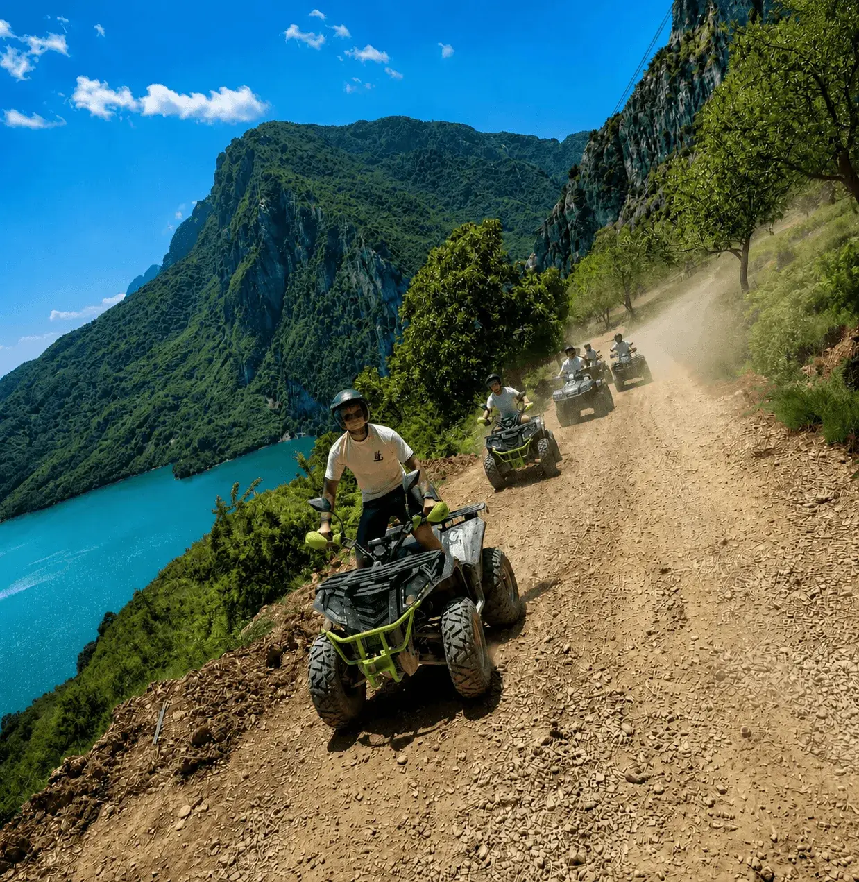 A line of riders on ATVs kicks up dust along a dirt mountain road above the turquoise waters of Lake Bovilla, with a wooden trail sign marking the route.