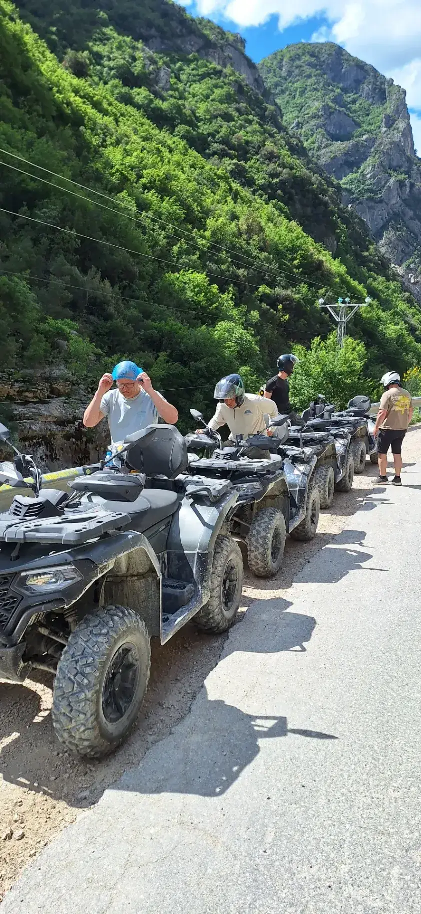 Riders fasten their helmets beside a line of parked CFMoto quad bikes on a mountain road below green cliffs, before the Bovilla ATV tour sets off.