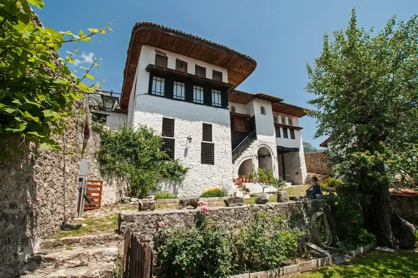 The Krujë Ethnographic Museum, a whitewashed Ottoman-era house with a deep wooden eave and dark shutters, seen from its terraced rose garden.