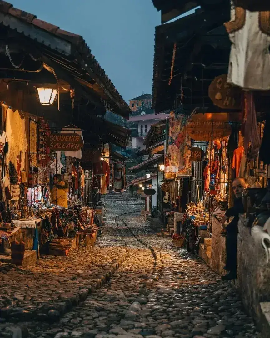 The cobbled lane of Krujë's Old Bazaar at dusk, lined with wooden shopfronts hung with carpets, textiles and souvenirs under warm lantern light.