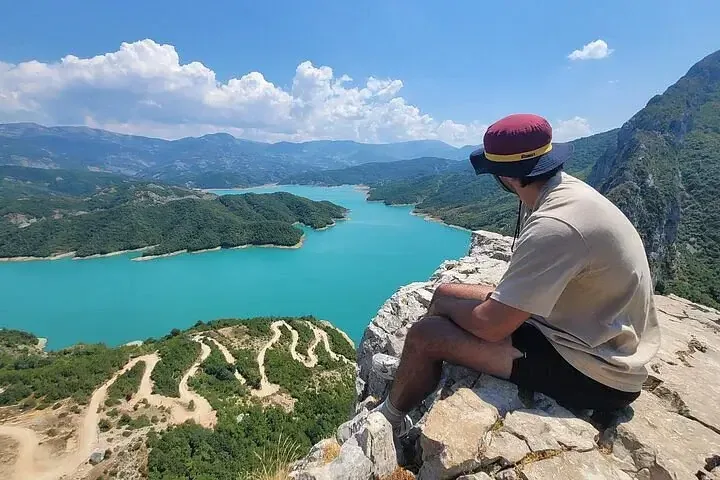 A hiker sits on a rocky outcrop above Lake Bovilla, looking out across the turquoise reservoir and the surrounding mountains.