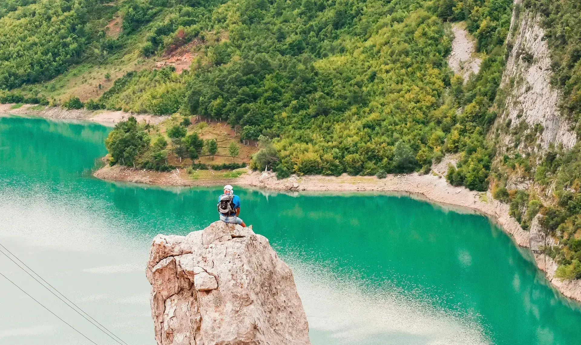 A hiker with a backpack sits alone on a limestone pinnacle high above the turquoise water of Lake Bovilla, with a forested ridge and pale cliffs behind.
