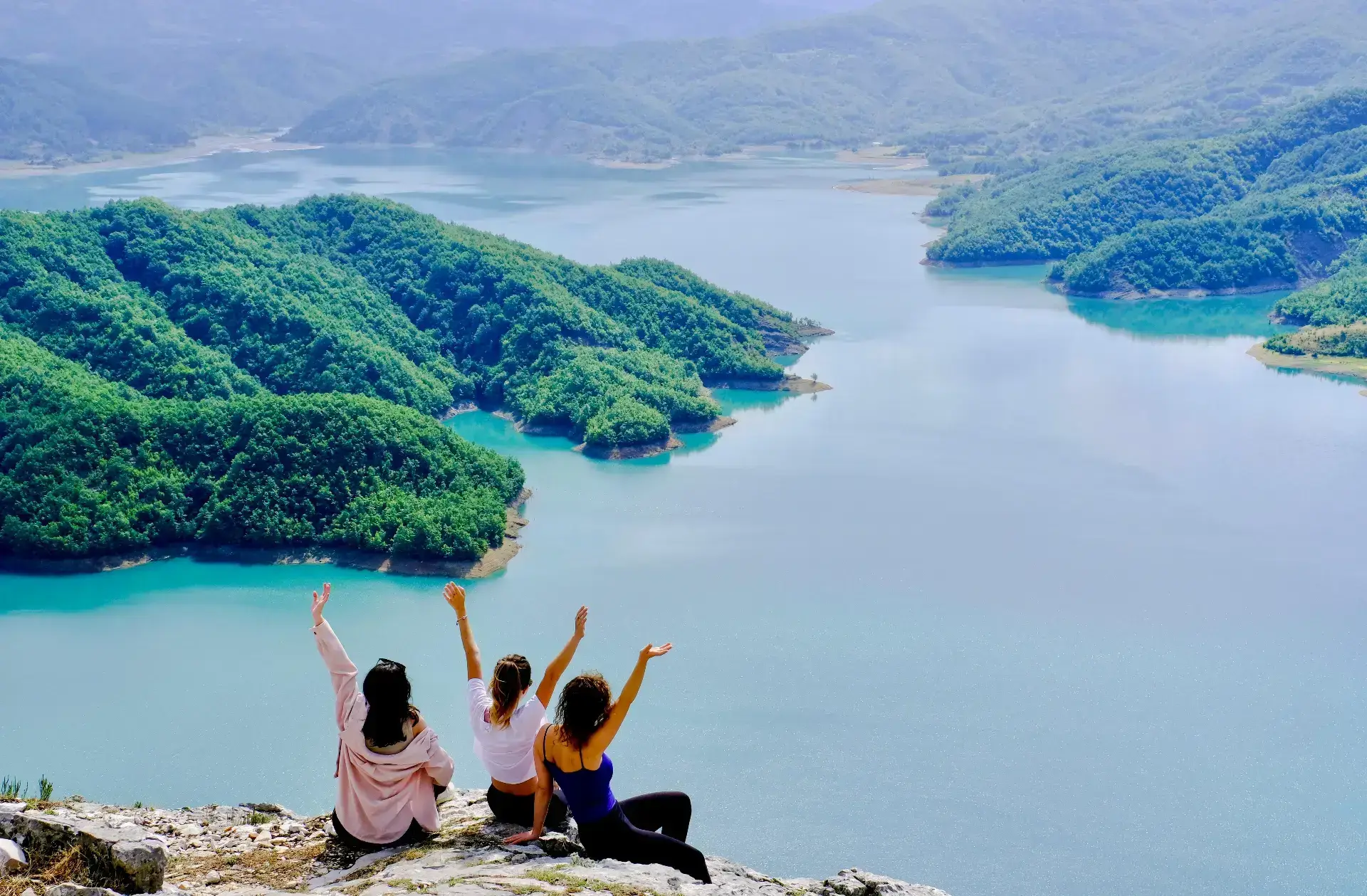Three hikers sit on the Mount Gamti clifftop with their arms raised, looking out over the forested inlets and islands of Lake Bovilla far below.