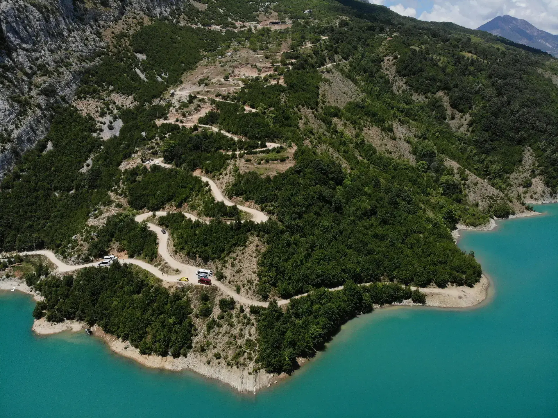 Aerial view of the hairpin gravel road winding down the forested mountainside to the turquoise shore of Lake Bovilla, with vehicles parked along it.
