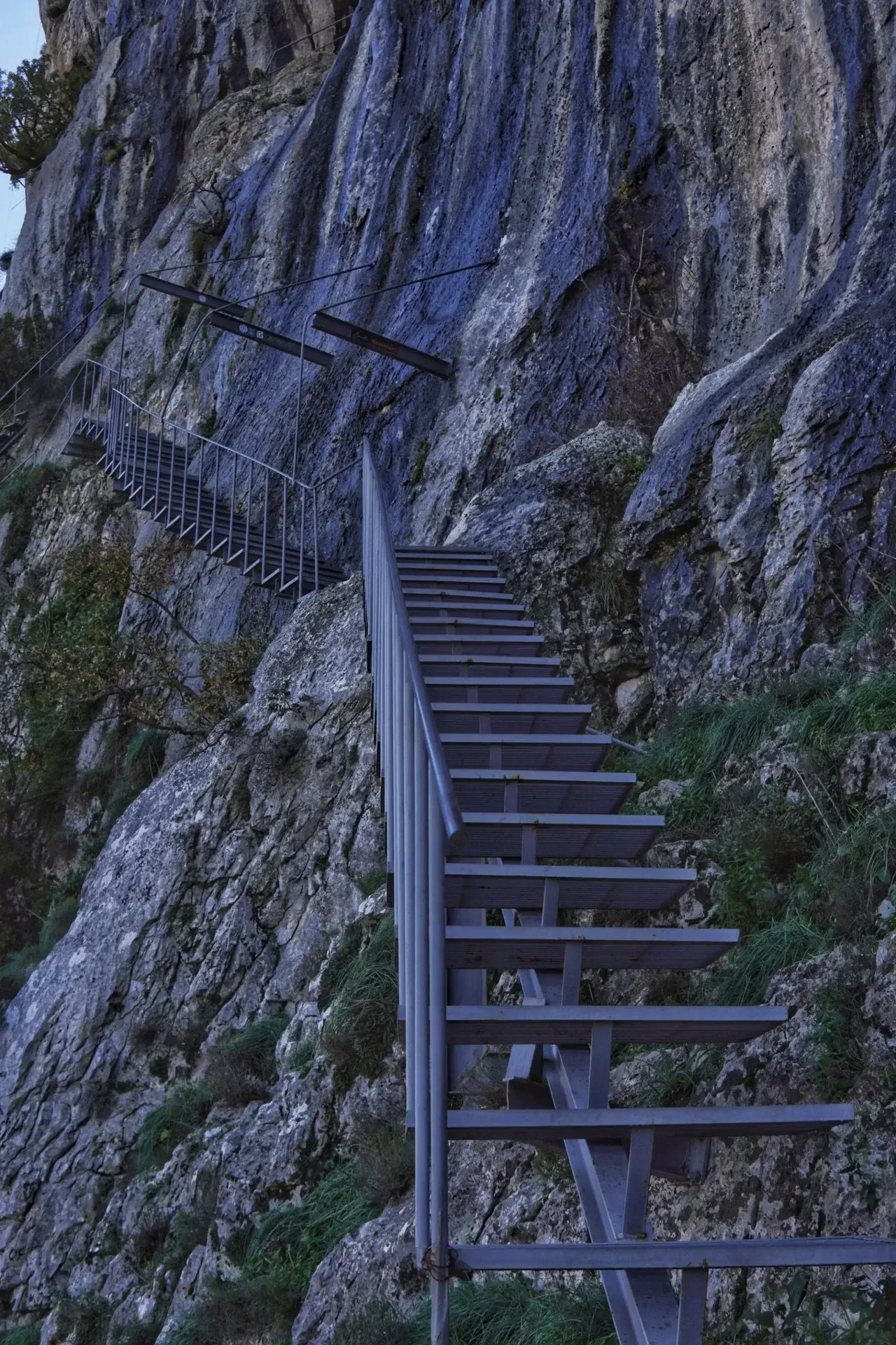 Steel staircase bolted into the limestone cliff face on the final approach to the Mount Gamti summit above Lake Bovilla.